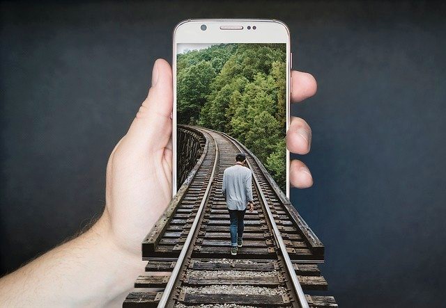 Hand holds mobile phone screen shows manipulated image of man walking along train tracks into mobile phone screen